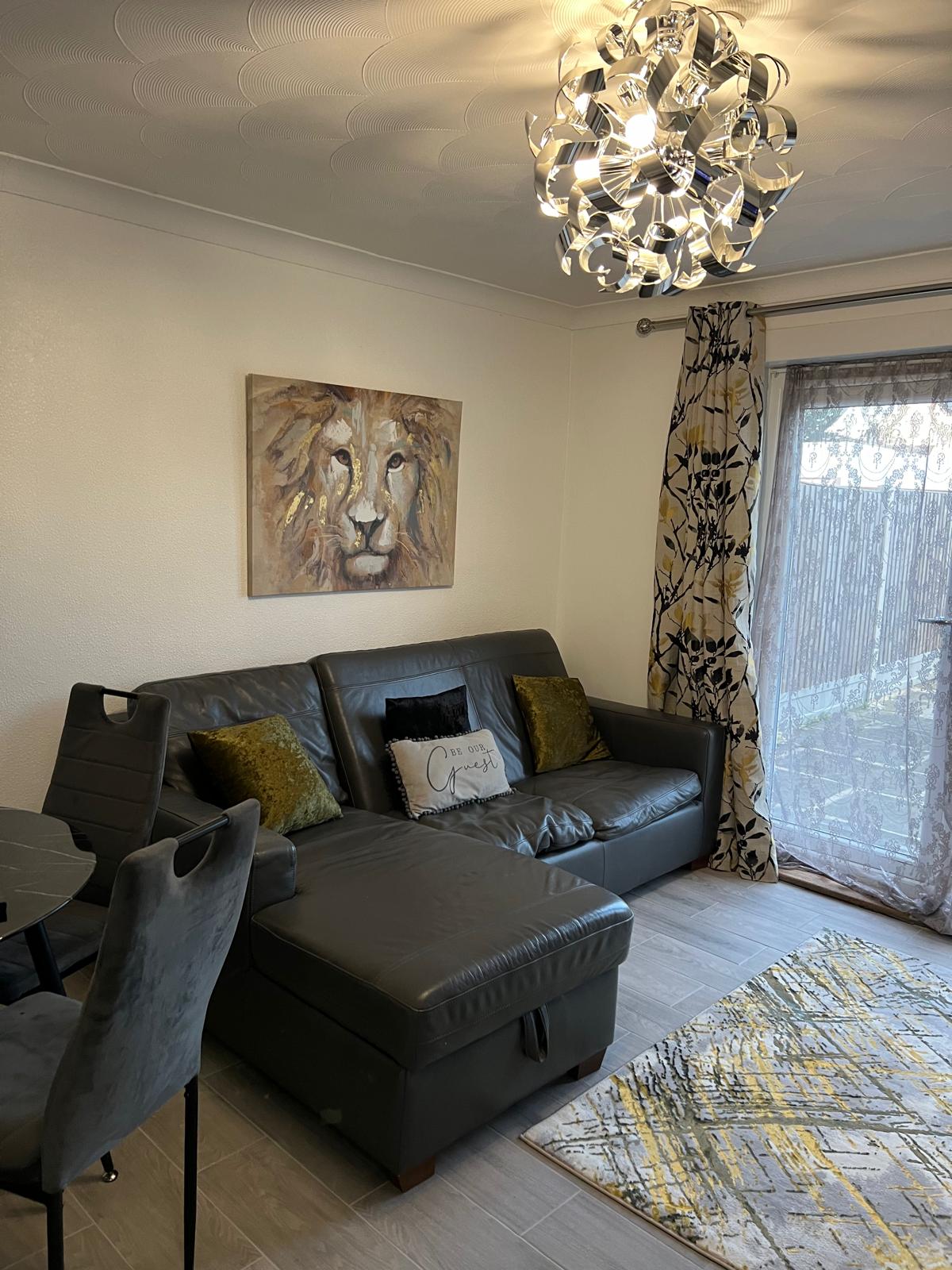 A contemporary living area is furnished with a dark leather sofa and a decorative accent pillow. A chandelier provides soft lighting, while a lion-themed artwork hangs on the wall. Additionally, a dining table for four is visible, with patterned curtains framing a glass door leading to the garden.