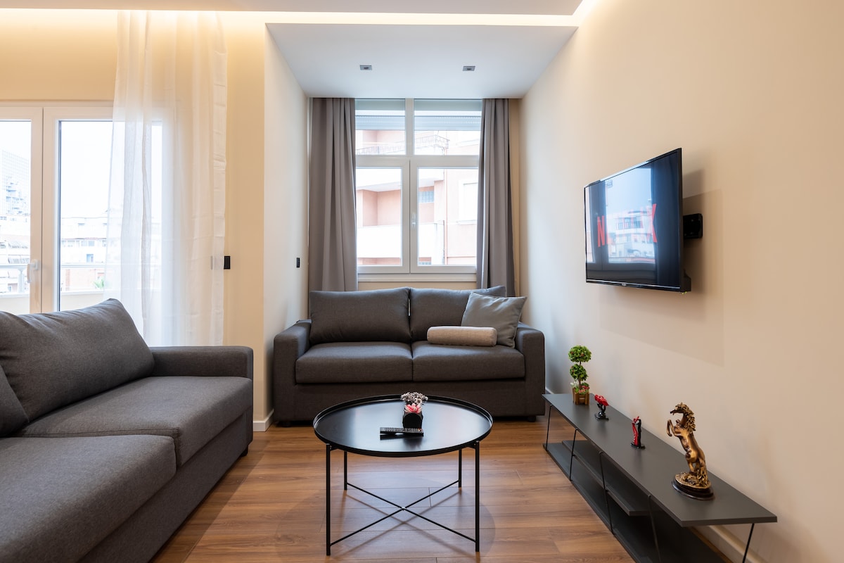 The living room features a spacious layout with two gray sofas arranged around a circular coffee table. A flat-screen TV is mounted on the wall, and natural light is provided by large windows, framed by soft curtains.