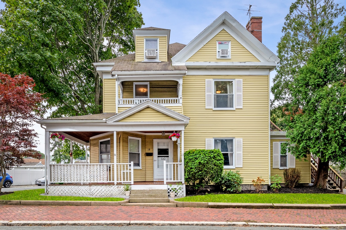 The exterior of a charming two-story yellow house is shown, featuring white trim and a welcoming front porch. Seasonal flowers hang from the porch railings. Lush greenery surrounds the property, complemented by a brick walkway leading to the entrance.