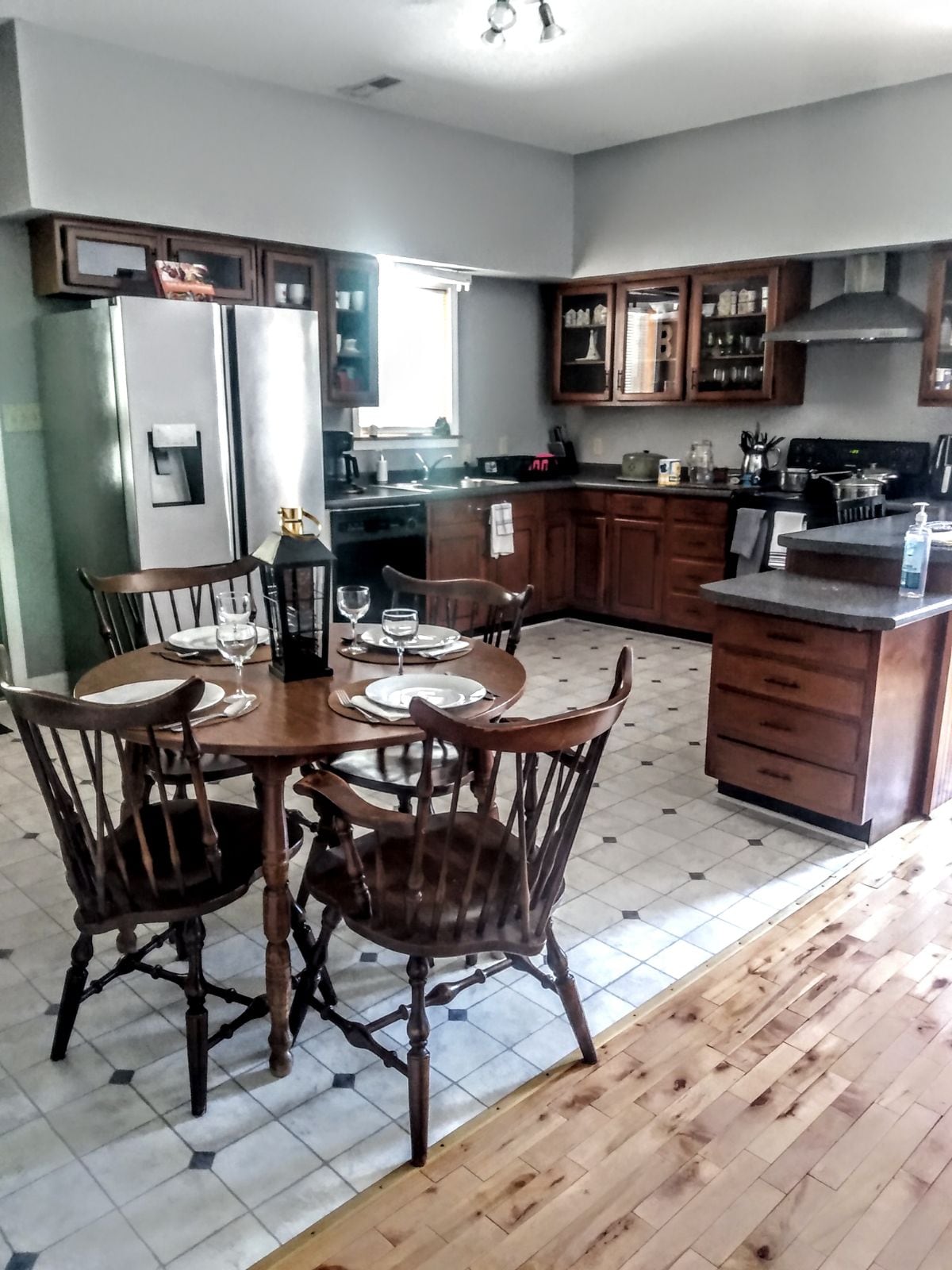 A spacious kitchen features wooden cabinetry and modern appliances, including a refrigerator and stove. A round dining table with four chairs is set for mealtime. The tiled floor complements the warm wooden accents throughout the room, creating a functional and inviting atmosphere.