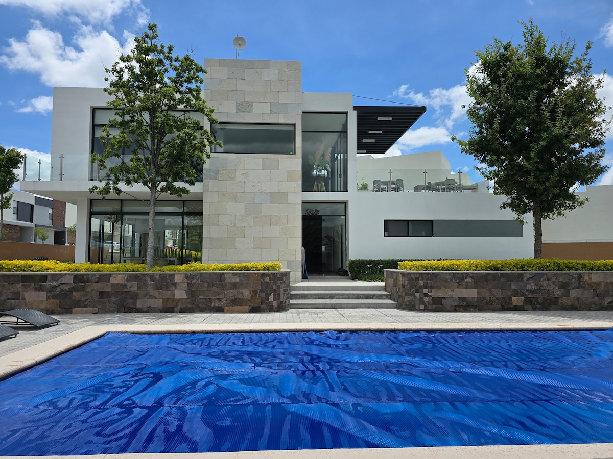 A modern home is showcased with a striking stone facade and large glass windows. A vibrant blue pool is located in the foreground, surrounded by well-maintained landscaping. Lush green trees frame the house, creating an inviting outdoor space.