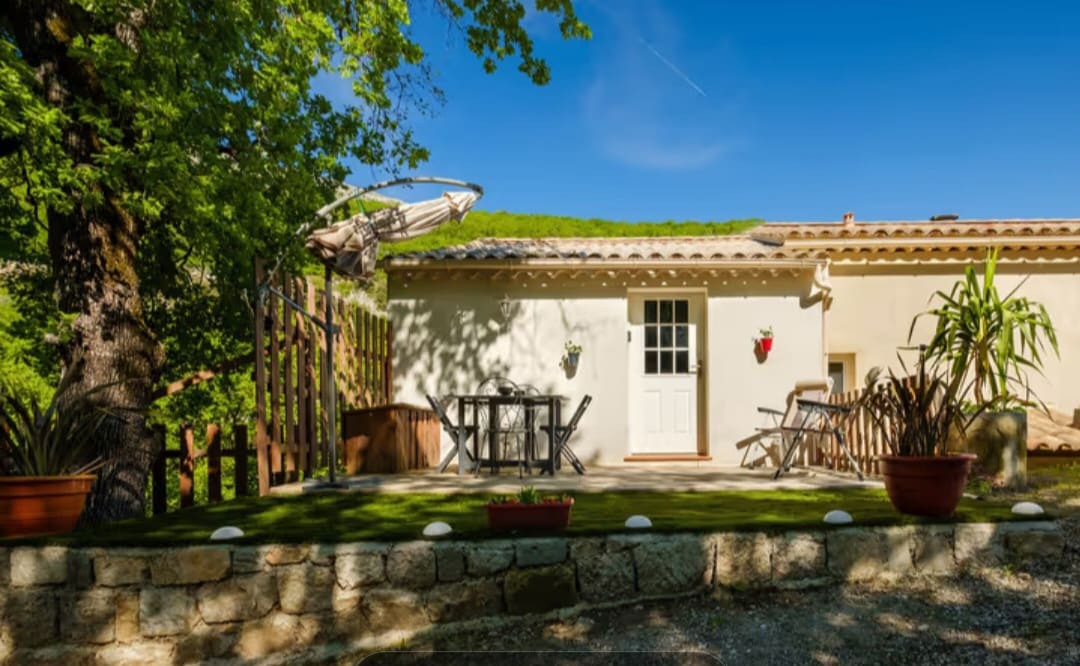 A charming terrace extends from the entrance of the studio, framed by potted plants. Light wooden furniture, including a table and chairs, is arranged for outdoor dining. A large tree provides shade on one side, enhancing the inviting atmosphere of the space.