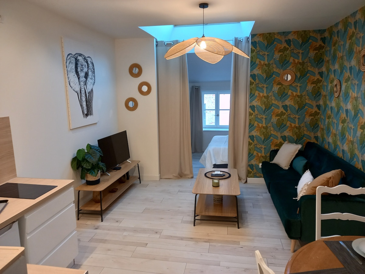 A living area is presented with a green sofa and a wooden coffee table. Light wood flooring is complemented by a colorful accent wall featuring a leaf pattern. A television is placed on a low cabinet. Natural light enters through a window behind sheer curtains, leading to a separate bedroom area.
