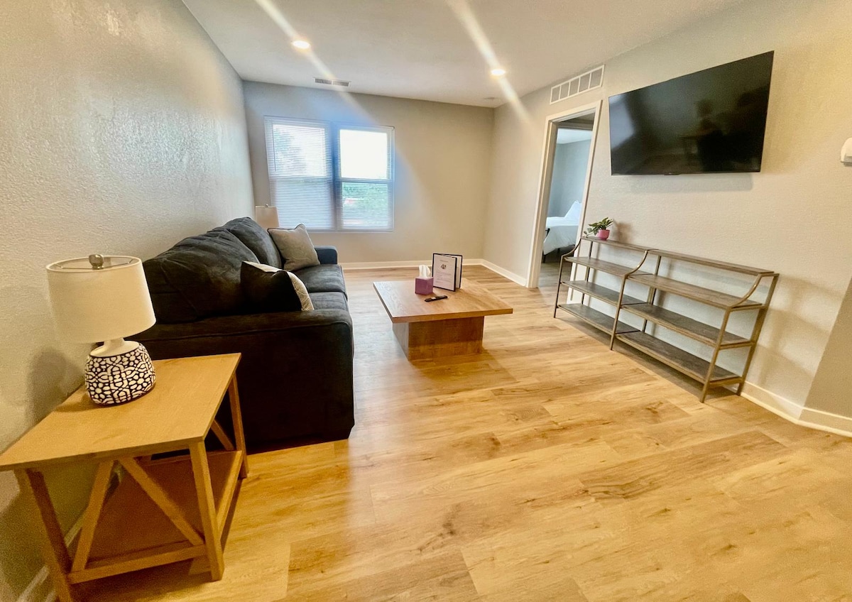 A well-lit living area features a comfortable dark sofa and a wooden coffee table. A decorative lamp sits on a side table. Natural light enters through two windows, highlighting the warm wood flooring. A wall-mounted TV is present, enhancing the space.