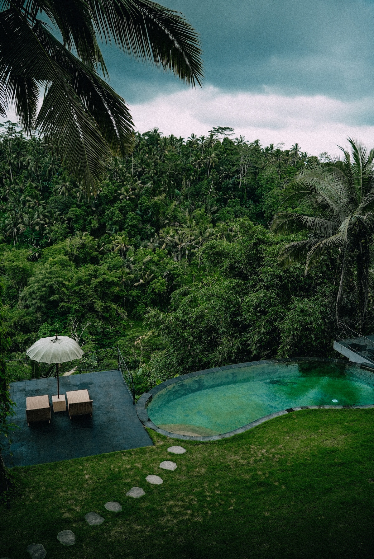 An infinity pool is set against a backdrop of lush green foliage, creating a serene retreat. Two loungers under an umbrella are positioned nearby, with a stone pathway leading to the pool area, surrounded by vibrant tropical scenery.