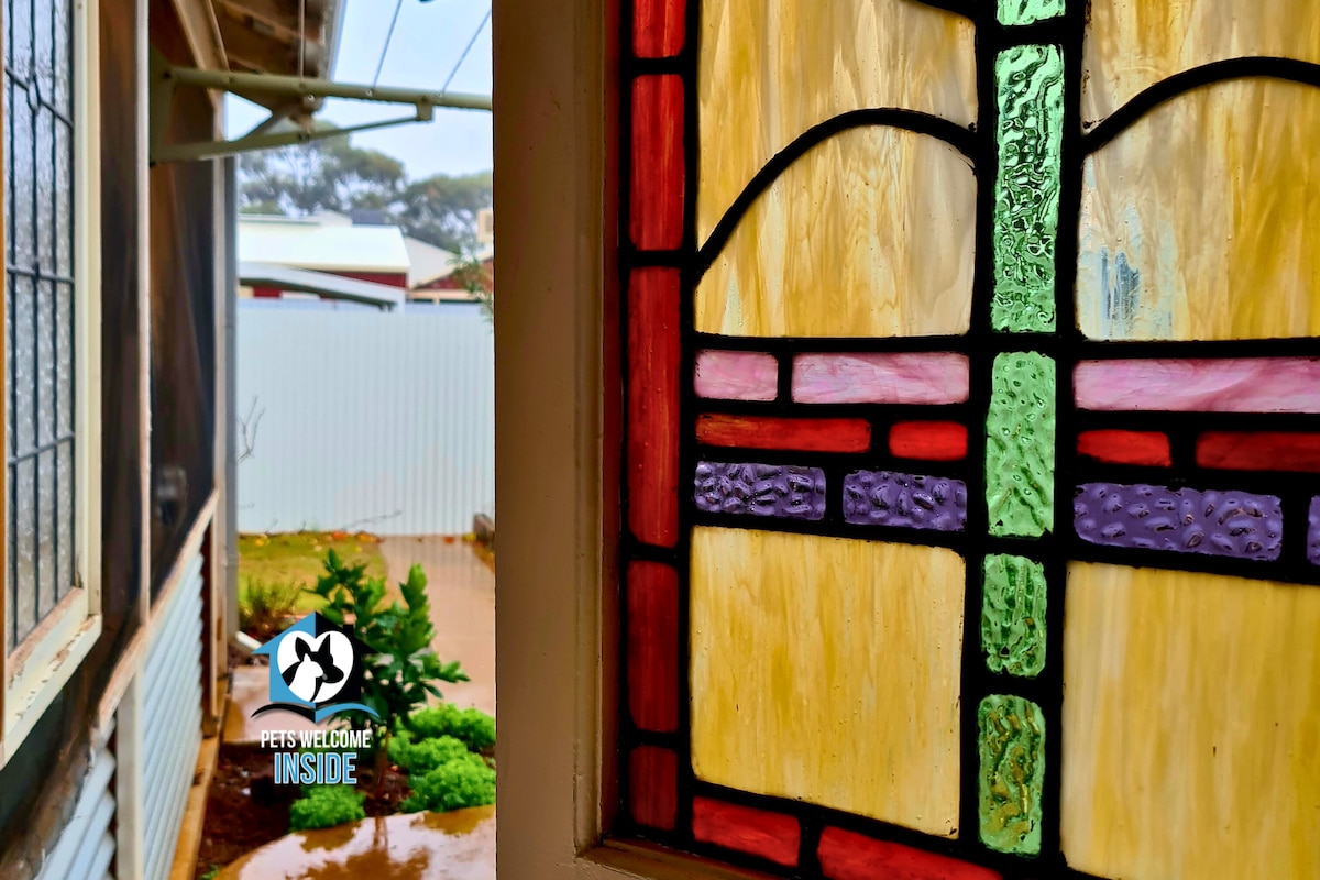 A close-up of a stained glass window displays colorful sections in hues of yellow, red, and green. Through the window, a glimpse of the backyard reveals greenery and a peeking shed, adding a sense of outdoor connection.