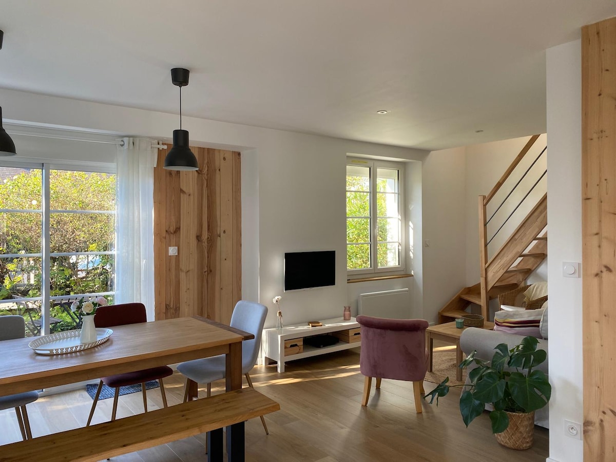 A light-filled open living area features a dining table with chairs, a cozy seating arrangement, and a television. Natural wood accents are present in the walls and staircase. Large windows allow ample sunlight, with greenery visible outside, creating a refreshing ambiance.