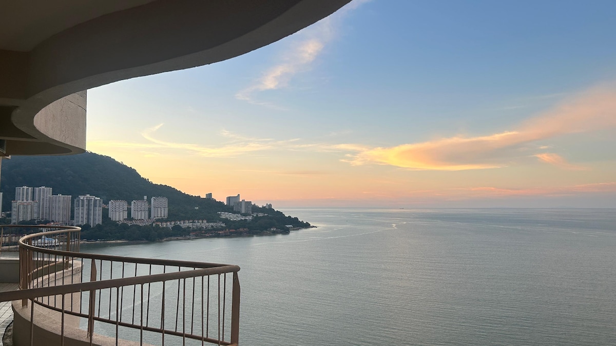 A panoramic view from a curved balcony, showcasing the tranquil sea meeting the horizon. Soft pastel colors fill the sky, reflecting on the water. Nearby coastal buildings and lush hills provide a backdrop to the serene landscape.