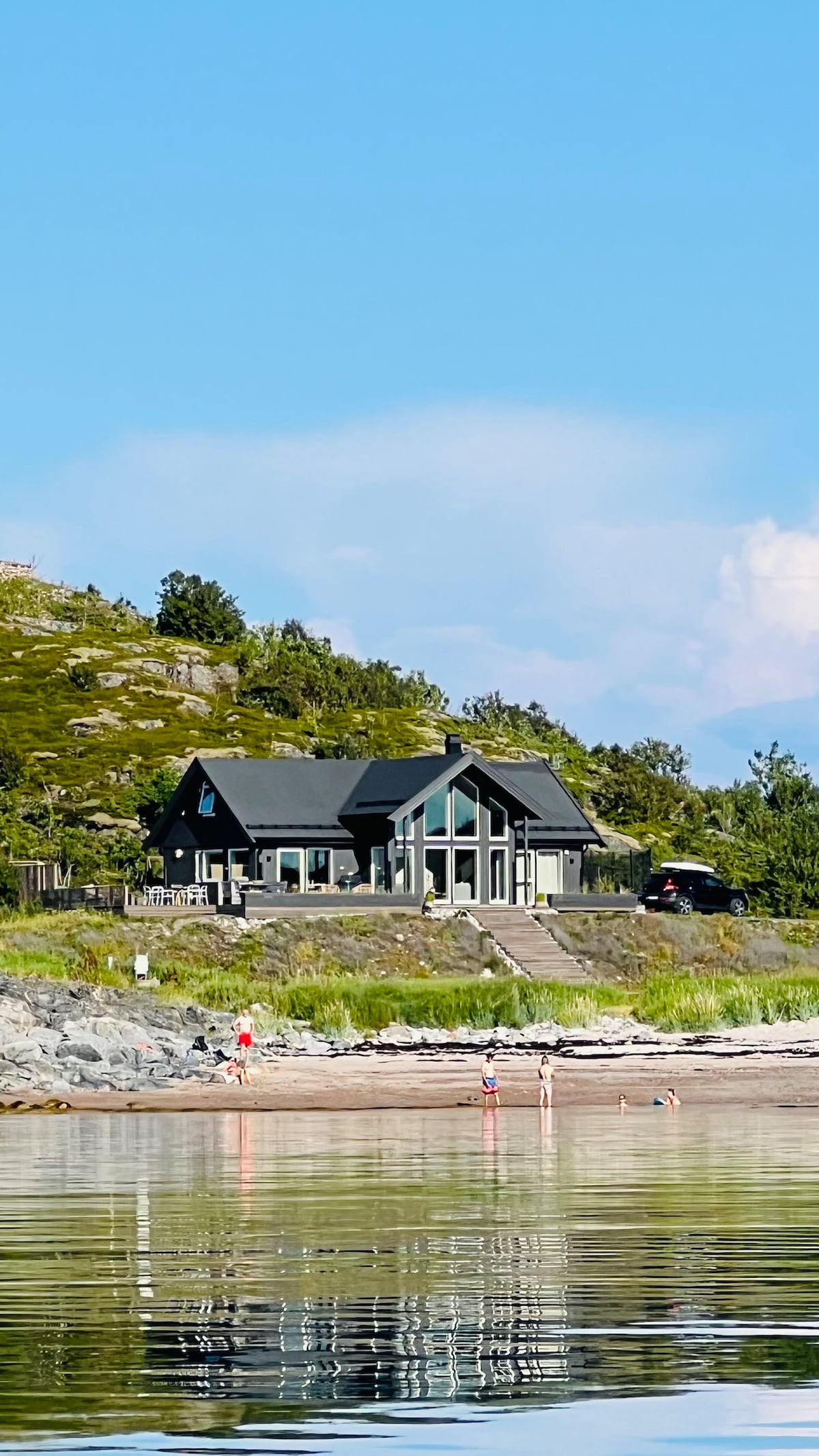 A modern cabin with a dark exterior is located near the shore. Large windows invite natural light and offer views of the surrounding landscape. The property is surrounded by greenery, and children can be seen playing in the water along the sandy beach.
