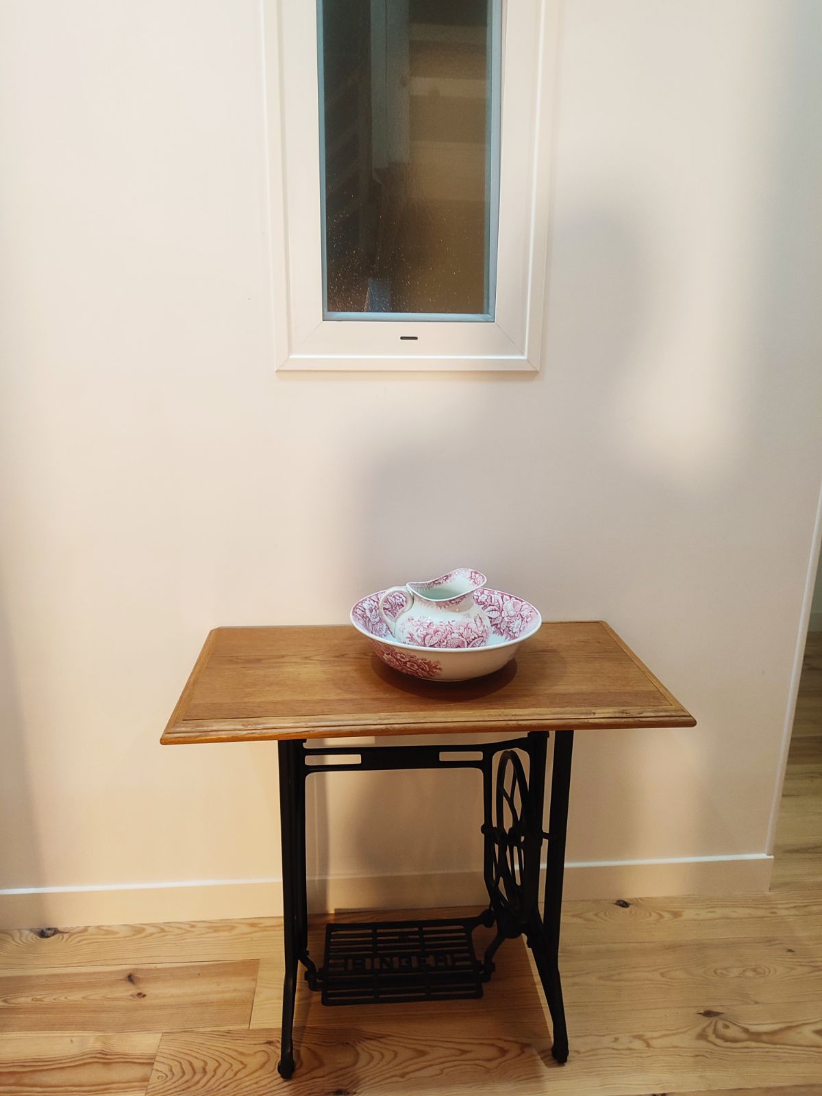 A vintage-style table with a dark frame and wooden top is seen against a light-colored wall. A decorative bowl with a floral pattern is featured prominently on the table, along with a small dish. A narrow window above allows natural light to enter the space.