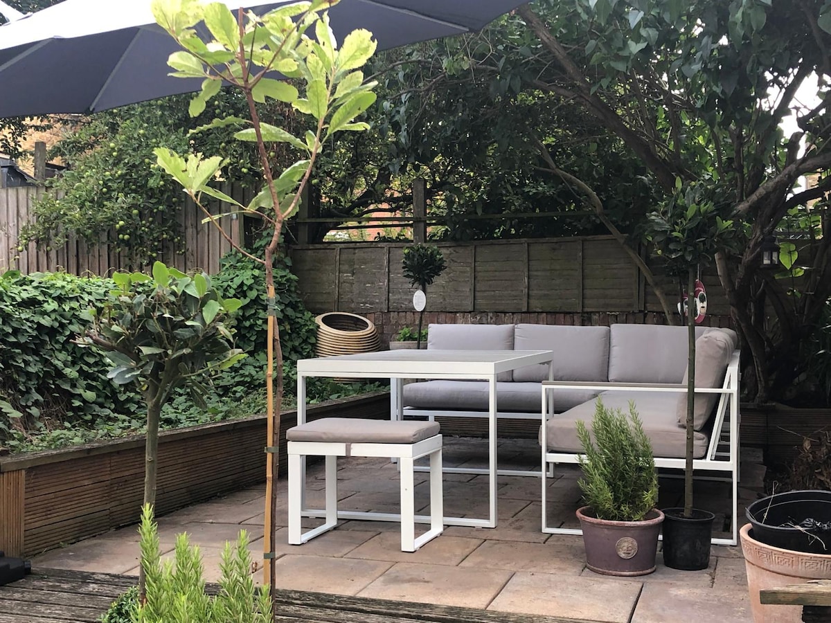 An outdoor garden area features a light-colored seating arrangement and a dining table, set on a stone patio. Lush greenery surrounds the space, complemented by potted plants. An umbrella provides shade, creating a welcoming environment for relaxation or outdoor meals.