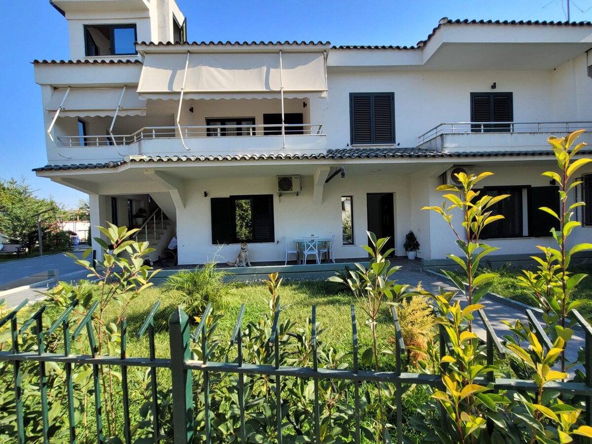 A two-story villa is set amidst a spacious garden, featuring a patio area with outdoor furniture. Lush green plants and grass surround the property, while a fence borders the front. The building displays a light exterior with multiple windows and a tiled roof.