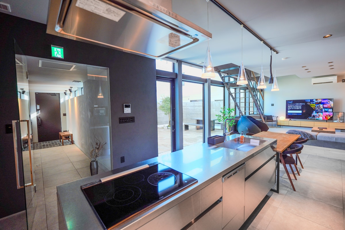 A modern kitchen features sleek stainless steel appliances and a spacious countertop. Through large windows, natural light illuminates the area. The open floor plan connects to a living space with a large TV visible in the background, promoting a welcoming atmosphere for social gatherings.