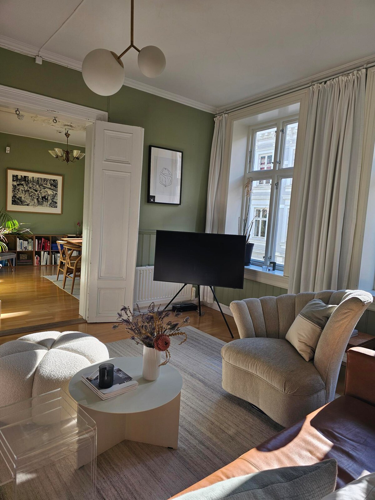 A well-lit living area features a plush armchair and a minimalist coffee table, accented by a vase of dried flowers. Large windows allow natural light to enter, revealing a view of the surroundings. A television is positioned nearby, with artwork displayed on the walls.