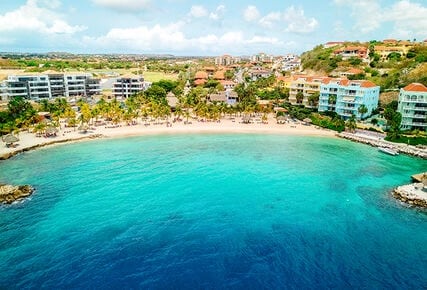 An aerial view captures a serene beach cove surrounded by palm trees, with gentle waves lapping at the shore. Colorful buildings are visible along the coastline, showcasing a blend of tropical architecture. The clear turquoise water creates a calming contrast with the sandy beach.