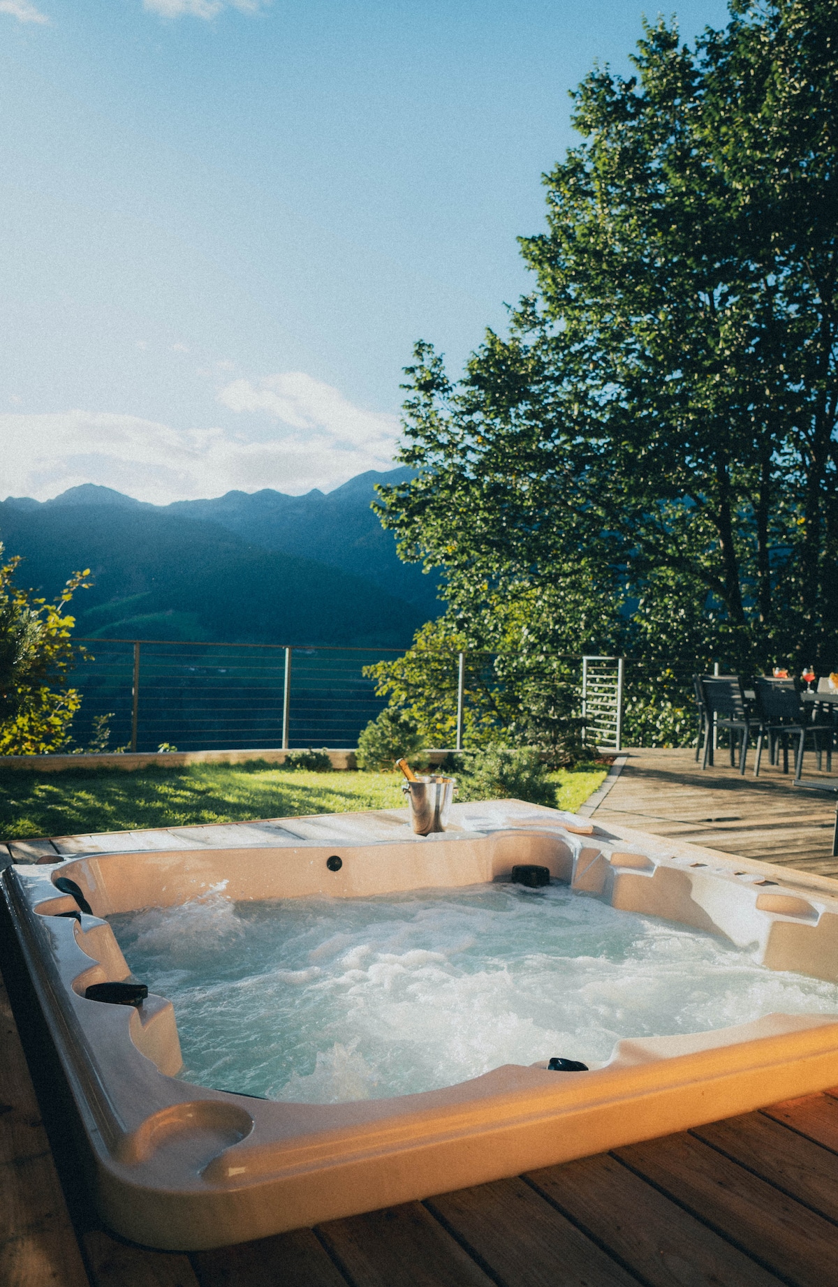 A private hot tub bubbles gently on a wooden terrace, surrounded by lush greenery. The backdrop features distant mountains under a clear blue sky, creating a serene outdoor space for relaxation.