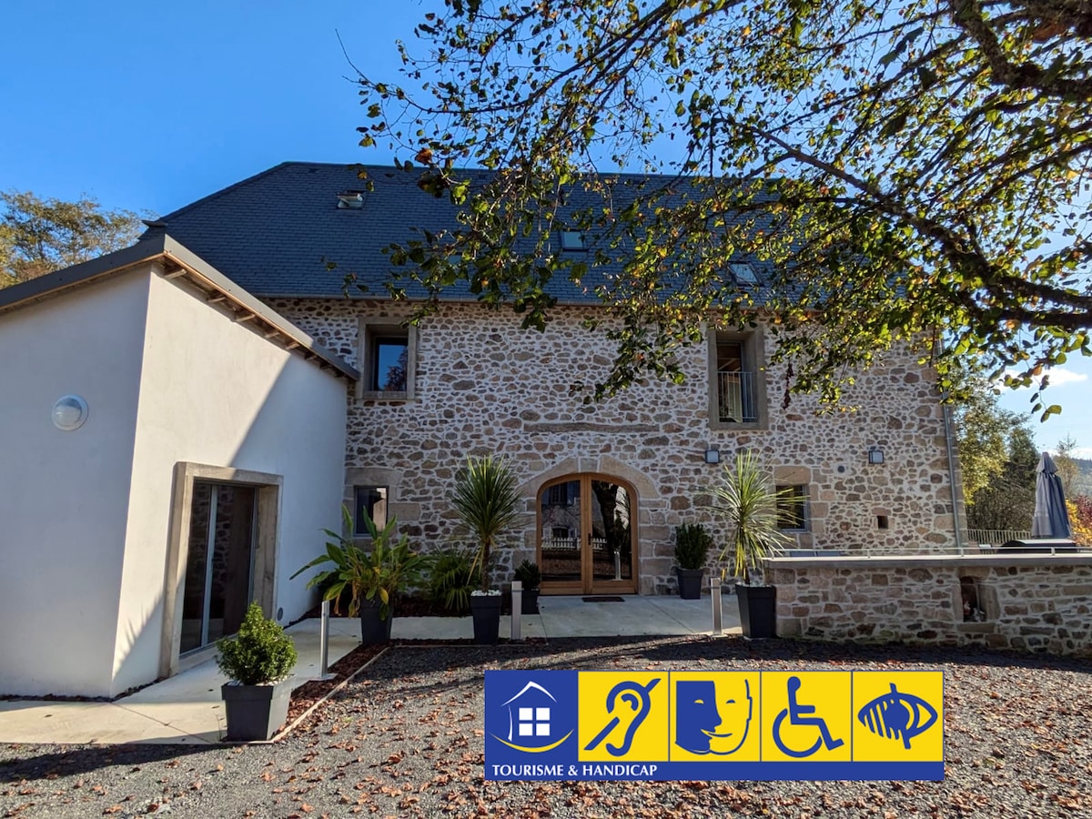 The exterior of a stone building is displayed, featuring wooden double doors and large windows. Potted plants line the entrance, and a textured gravel driveway leads to the entrance. Assurances of accessibility are represented by visual symbols of inclusivity at the bottom.