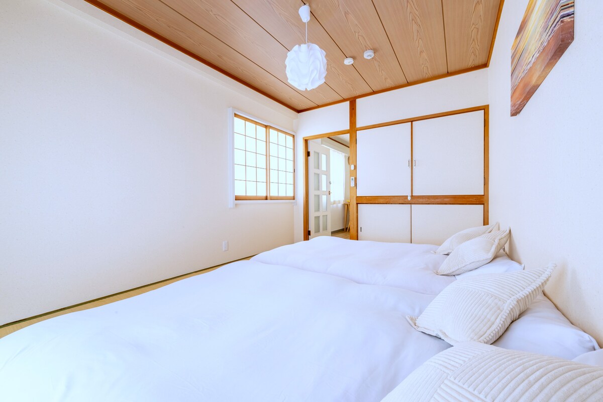 A spacious room featuring a double bed with crisp white bedding and decorative cushions. Natural light enters through a window adorned with a grid pattern. Neutral tones and wooden accents create a calming environment, while sliding doors provide access to other areas.