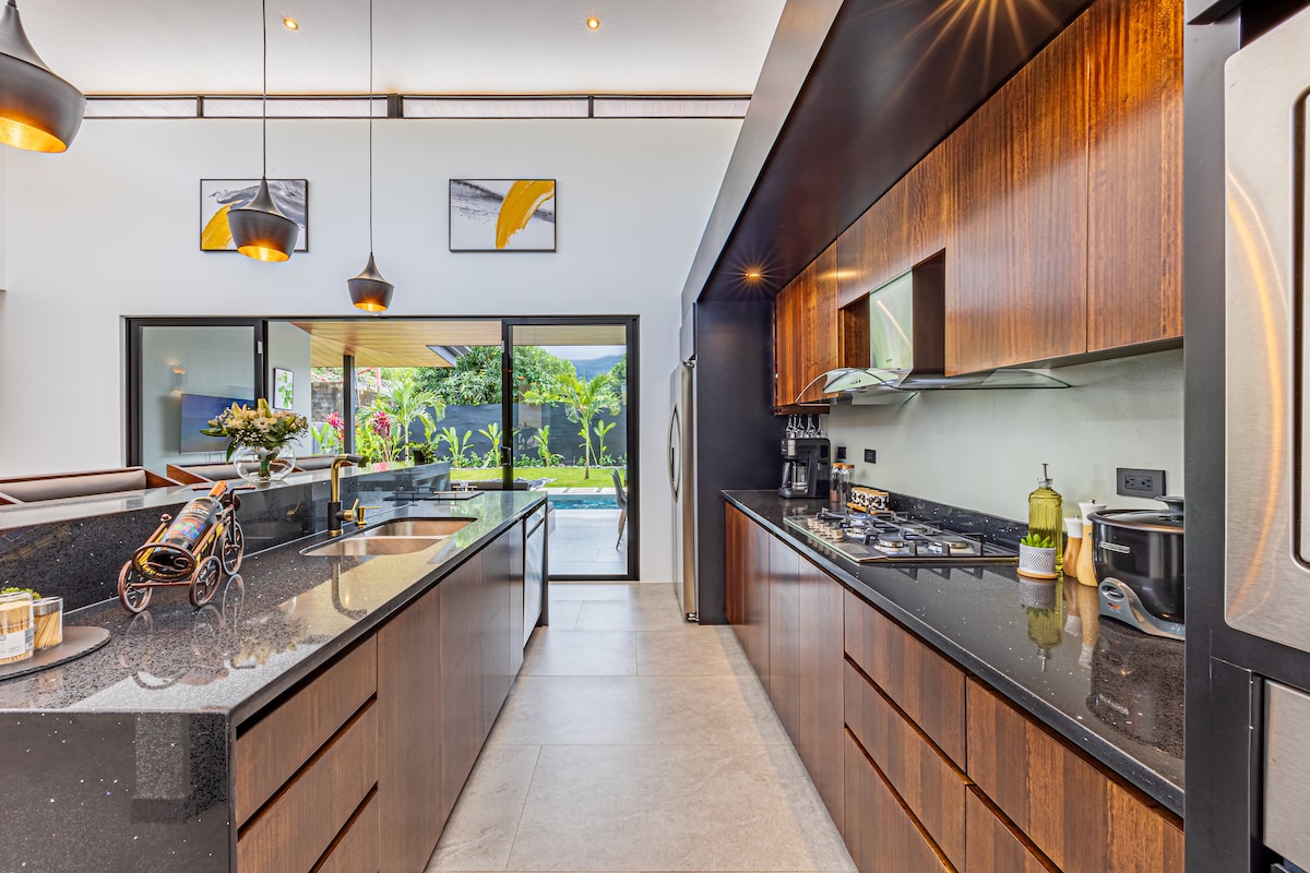 A modern kitchen features sleek dark cabinetry and a spacious layout, complemented by granite countertops. Large windows offer a view of the backyard, showcasing greenery and the private pool. Stainless steel appliances are neatly integrated, enhancing the kitchen's functionality.