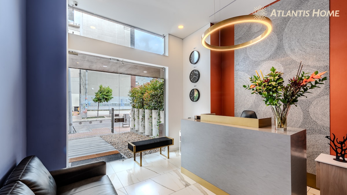 The reception area features a modern front desk with a sleek design. Natural light floods the space through large glass doors, which open to an outdoor area. A black sofa is positioned along one wall, complemented by decorative plants and contemporary artwork.