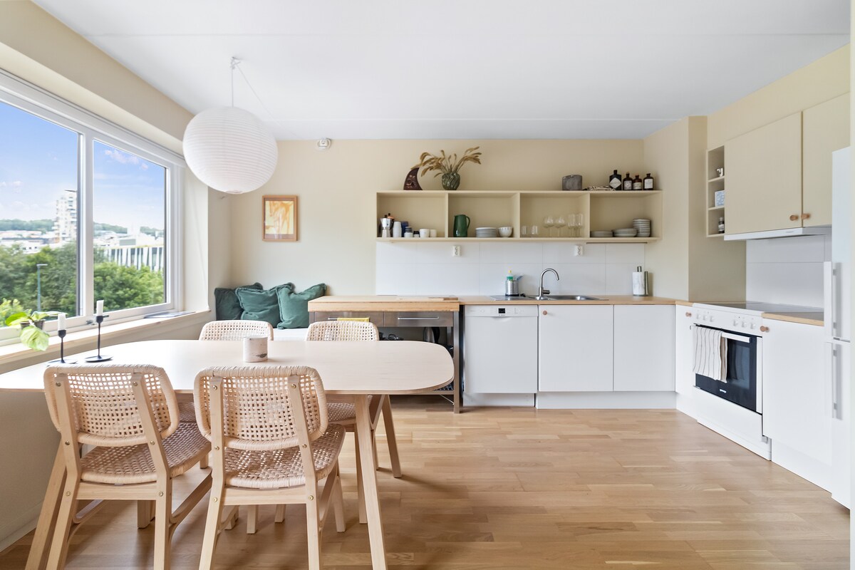 A modern kitchen and dining area feature a light wooden table surrounded by four wicker chairs. Open shelving displays dishware, while the kitchen includes appliances and ample counter space. Large windows offer views of greenery, allowing natural light to illuminate the space.