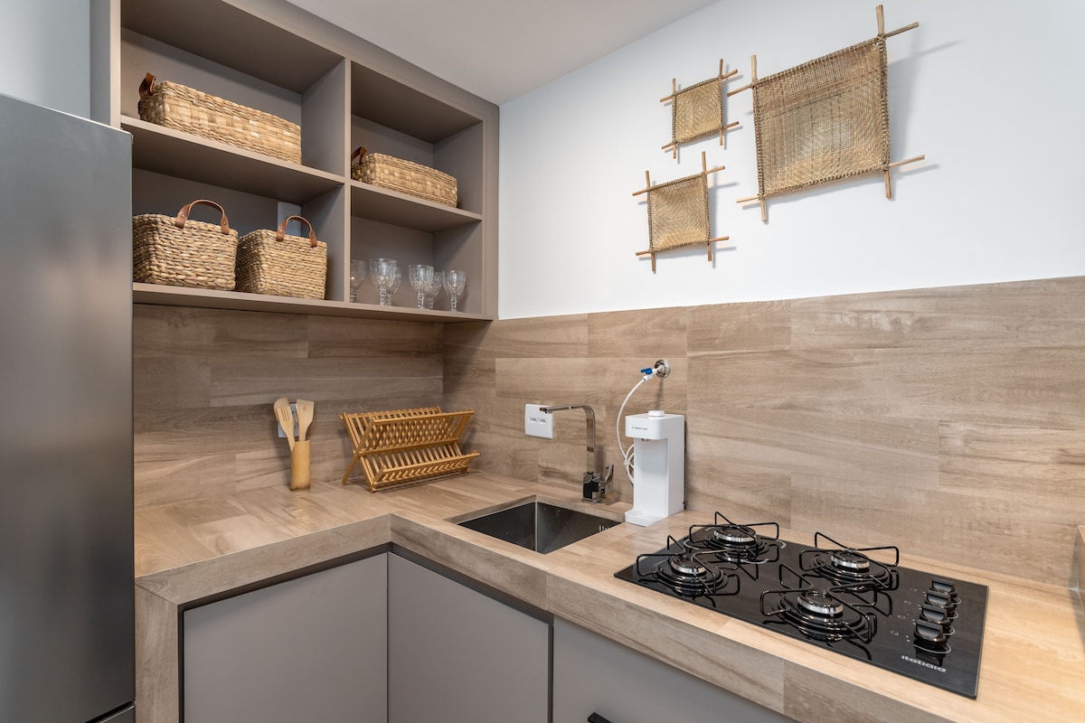 The image showcases a modern kitchen with a light wood finish. Countertops are complemented by grey cabinetry and open shelving displaying woven baskets and glassware. A stainless-steel sink is positioned beside a four-burner gas stove, creating a functional cooking space.