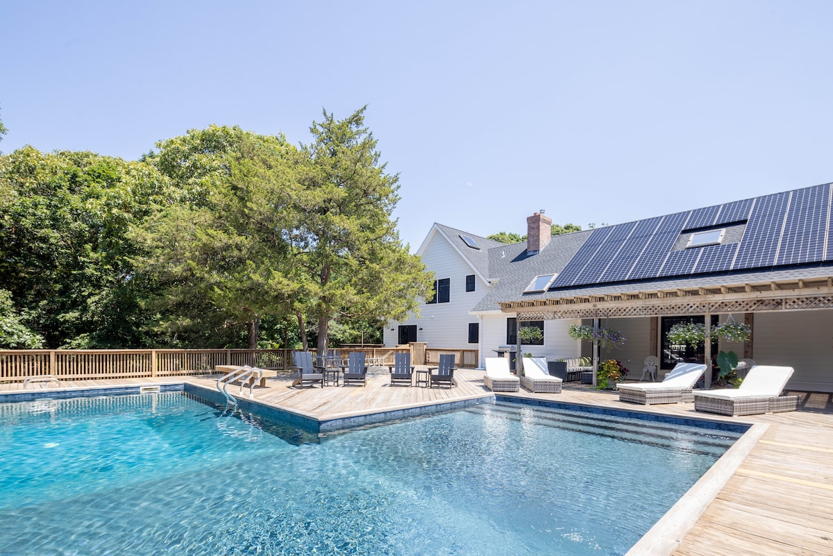 A large L-shaped pool is surrounded by a wooden deck, equipped with lounge chairs and shaded seating areas. The modern farmhouse, featuring a sloped roof, is situated nearby among lush greenery, creating a serene outdoor environment for relaxation and enjoyment.