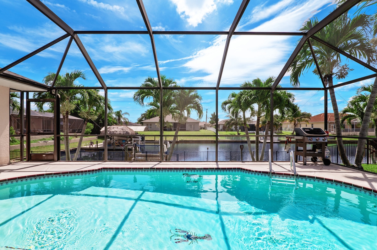 The outdoor space features a heated pool surrounded by a screened enclosure, providing unobstructed views of the canal and palm trees. A barbecue grill is situated nearby, offering options for outdoor dining and relaxation under a clear blue sky.