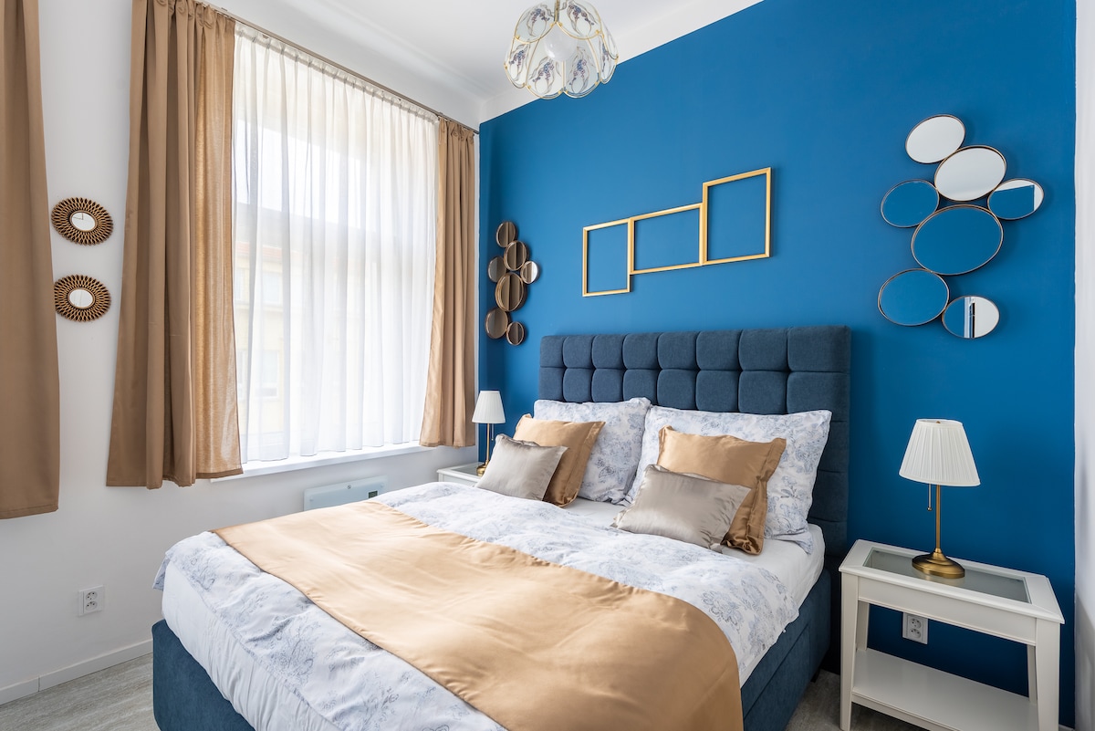 A bedroom features a spacious bed with a plush blue headboard, adorned with decorative pillows. The walls are painted in a calming blue, complementing the warm beige bedding. A large window is dressed with light curtains, allowing natural light to fill the room, and two bedside lamps are positioned on either side.