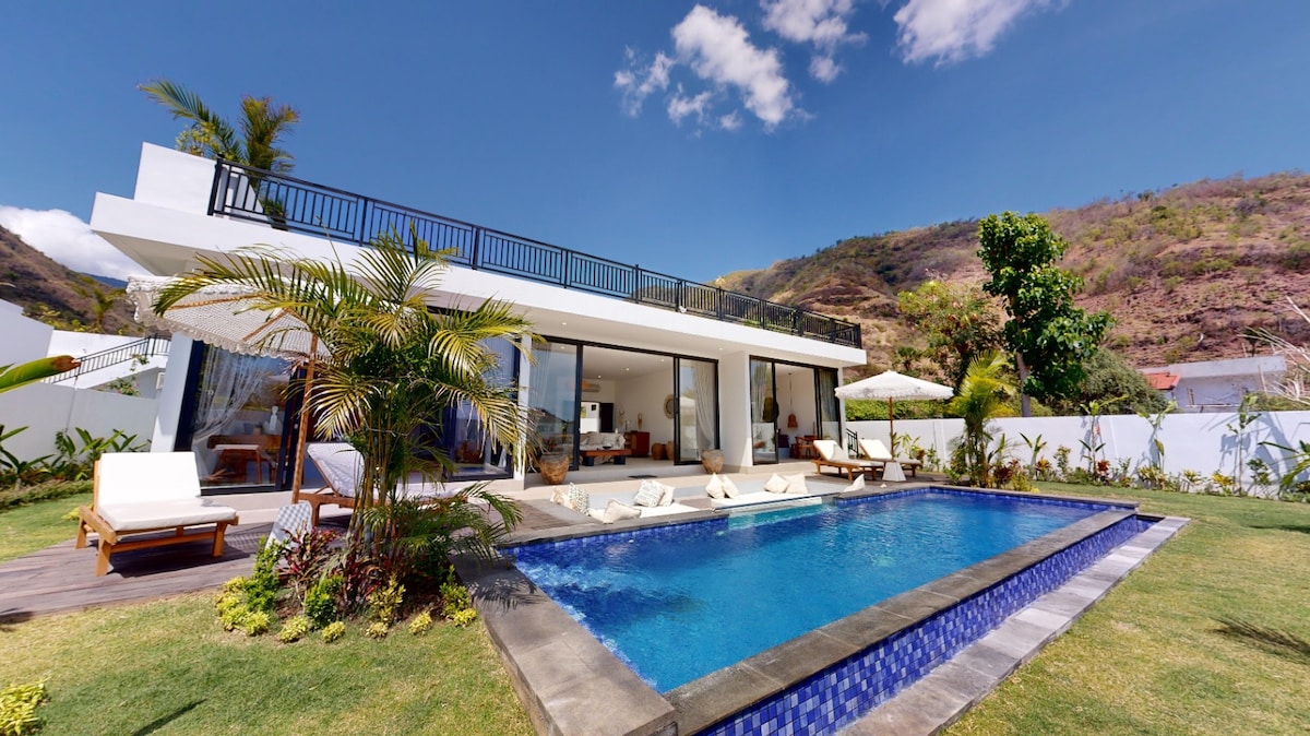 A modern villa is showcased with large glass doors opening to a private pool area. Lush greenery and tropical plants surround the pool, which is adorned with lounge chairs. The landscape features gentle hills in the background, indicating a tranquil setting.