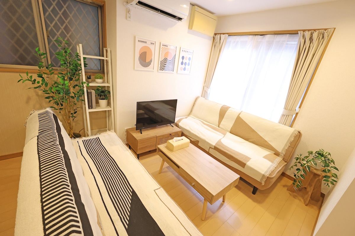 A modern living room is featured, equipped with a light wooden coffee table and a low-profile TV unit. Two comfortable sofas are arranged to encourage conversation, complemented by framed artwork on the walls. Natural light filters in through sheer curtains, enhancing the space's fresh ambiance.