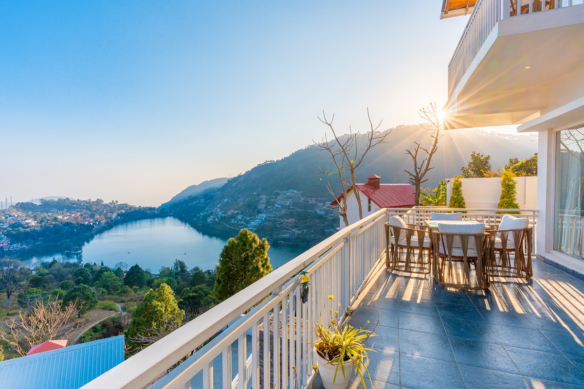 A spacious balcony offers a dining table surrounded by chairs, overlooking a serene lake and lush hills. Soft sunlight filters through trees, creating a calming ambiance. The view captures the beauty of nature, providing an inviting space for relaxation and enjoyment.