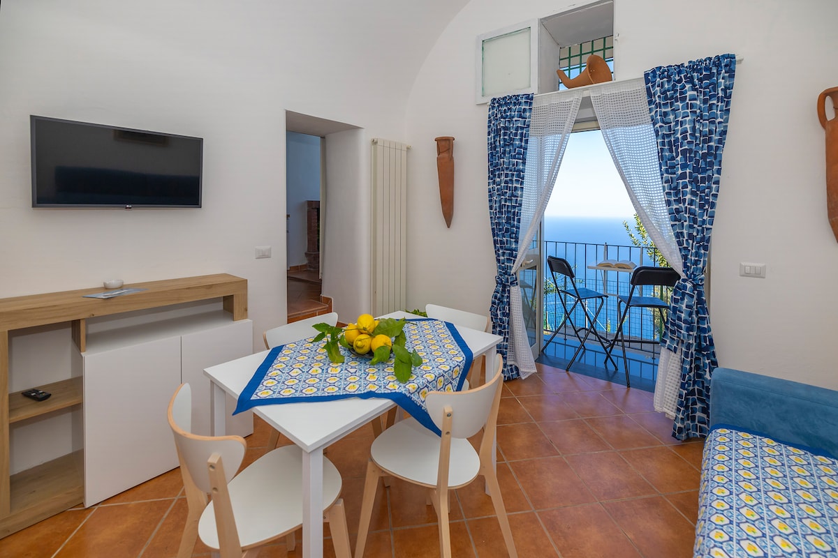 A dining area is depicted with a white table set for four, surrounded by light wooden chairs. Bright blue curtains frame a window that offers views of the sea, complemented by decorative pottery on the wall. A television is mounted above a light wood cabinet.
