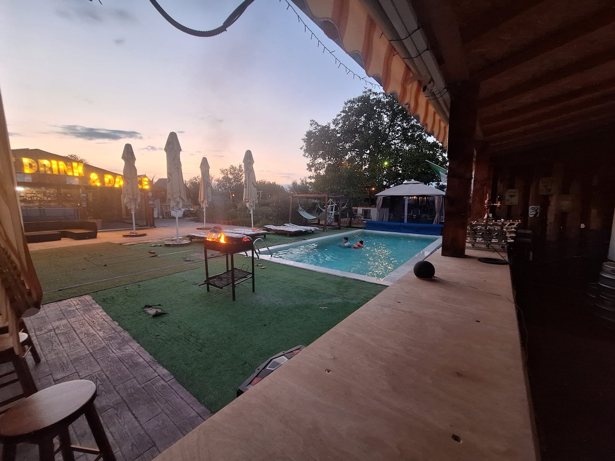 An outdoor space features a private swimming pool, surrounded by a turf area. A barbecue grill is visible, with flames flickering above. Sun loungers and umbrellas line the poolside, while a few trees provide greenery. The setting is illuminated by the soft light of dusk.
