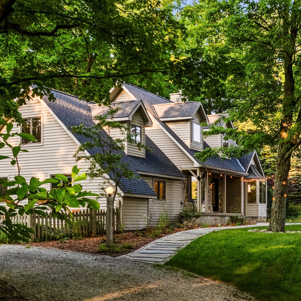 A charming house is nestled among lush greenery, featuring a front porch with columns and several windows that invite natural light. A gravel path leads to the entrance, surrounded by neatly landscaped gardens and trees, enhancing the peaceful rural ambiance.
