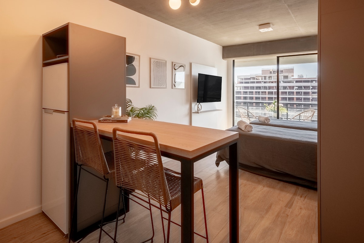 A modern studio apartment features a functional layout with a small dining area and three bar stools. A wall-mounted TV is positioned above a console. Natural light is welcomed through a large window, offering views of the building's exterior.
