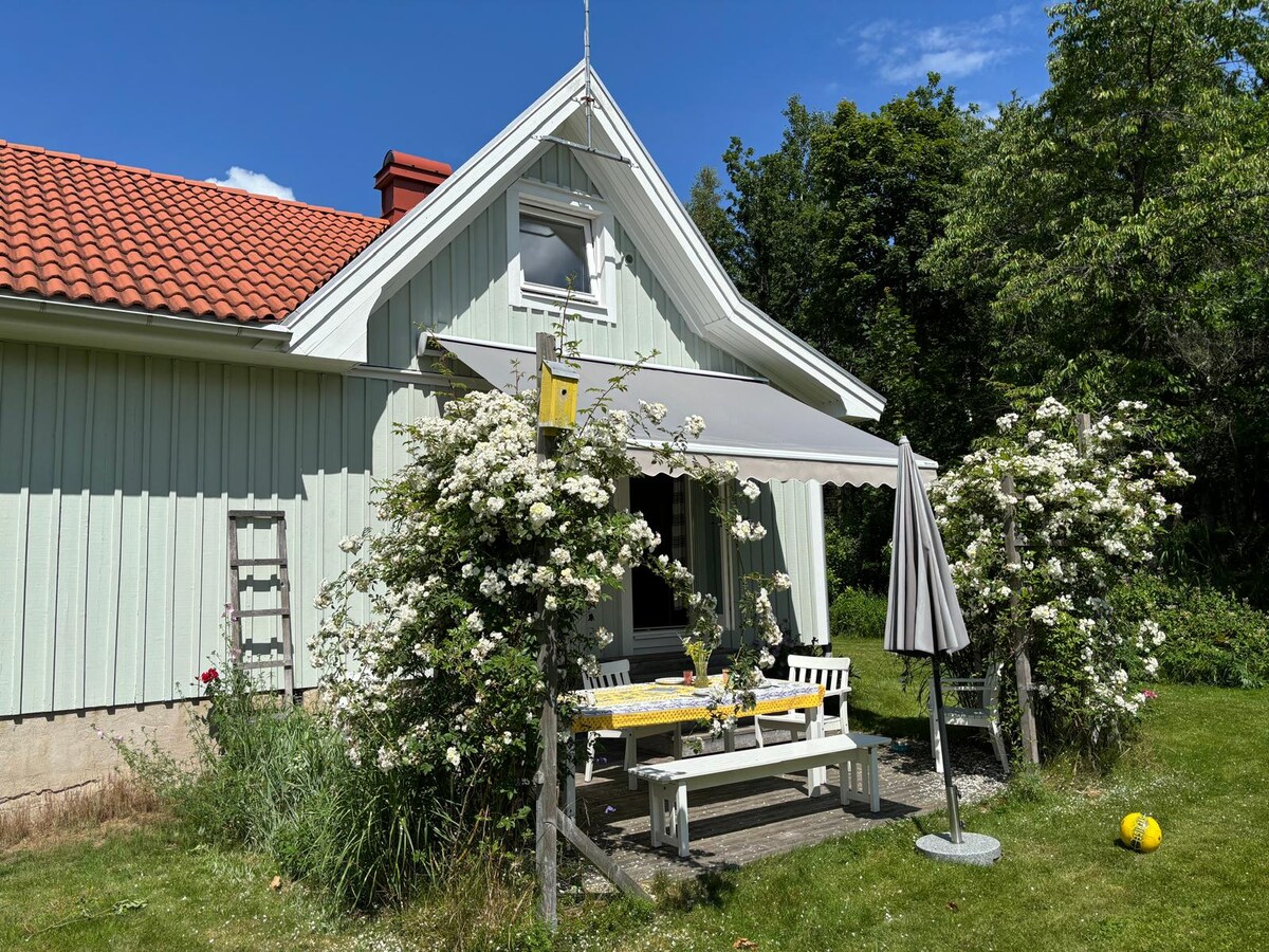 A charming light-green house displays a welcoming facade, accented by a red-tiled roof. White outdoor furniture offers a seating area beneath a large awning, surrounded by lush greenery and blooming flowers. A decorative birdhouse adds an inviting touch to the garden view.