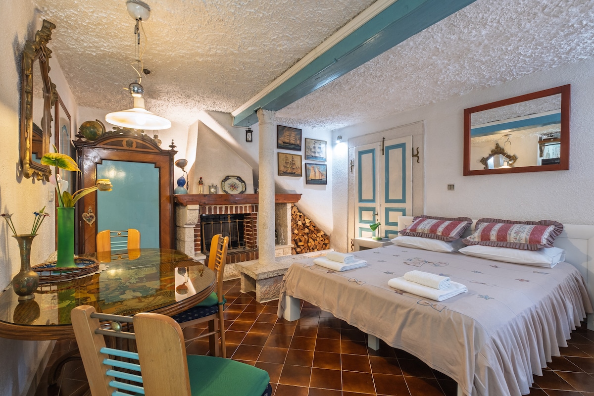 A cozy living space features a spacious bed with neutral bedding and decorative pillows. A dining table with four chairs is nearby, and a traditional fireplace is present, complemented by logs stacked beside it. An accented blue beam runs along the ceiling, enhancing the room's character.