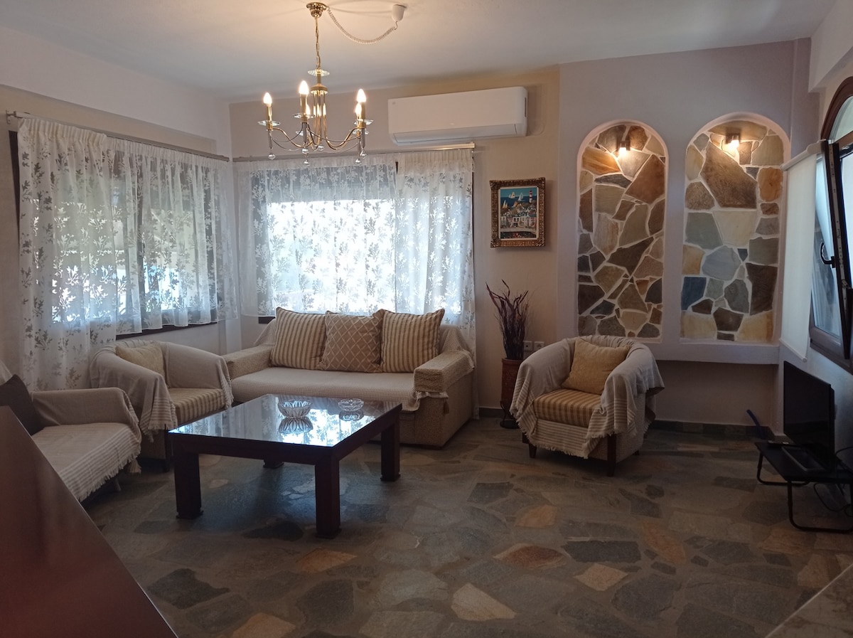 A cozy living area is furnished with a spacious sofa and two armchairs, complemented by decorative throw blankets. Natural light filters through large curtain-covered windows, while a coffee table occupies the center of the room. A stone feature wall adds texture to the space.