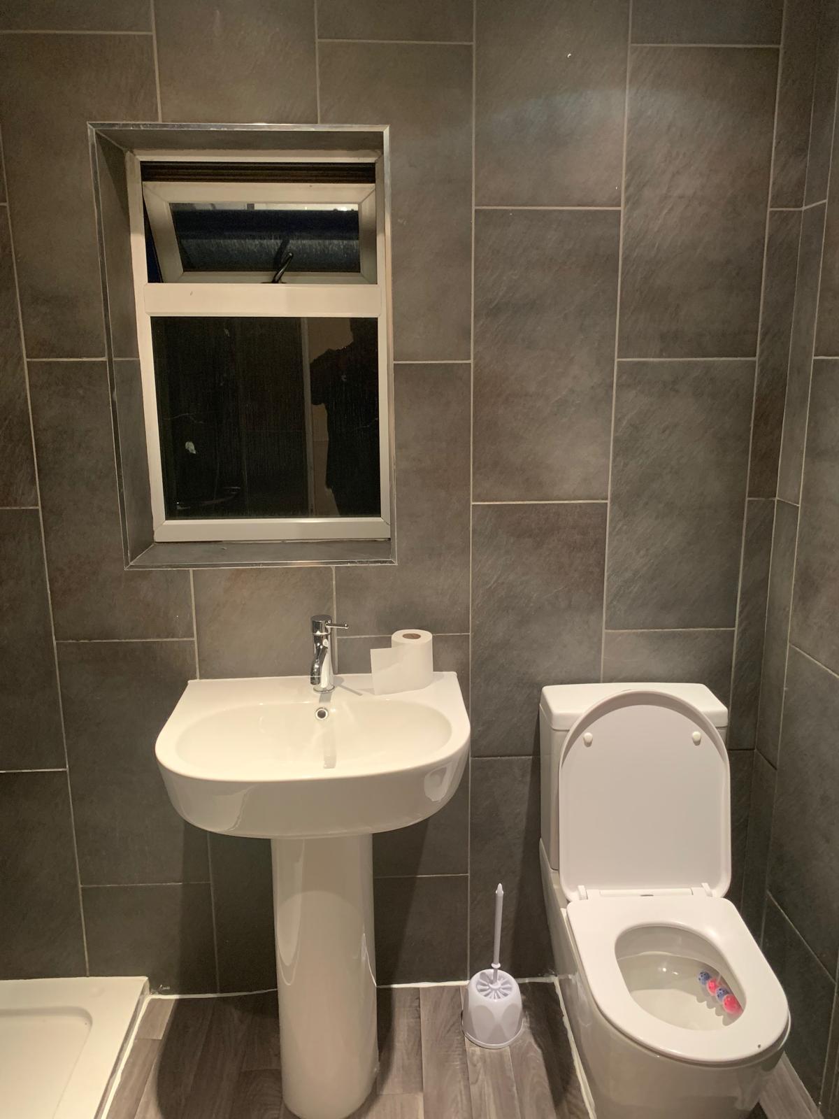 A modern bathroom features sleek gray tile walls and flooring, complemented by a white pedestal sink and toilet. A window allows natural light to enter, enhancing the clean and spacious feel of the room. Essential toiletries are placed on the sink.