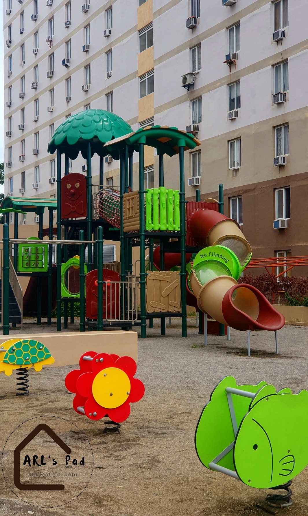 A vibrant playground area features various play structures, including slides and climbing equipment. Brightly colored elements, such as a flower and fish, are present on the ground, providing a playful environment. Surrounding buildings are partially visible, emphasizing the accessibility of outdoor recreational space.