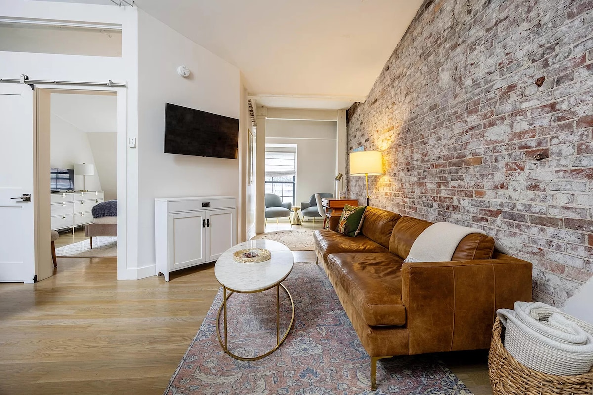 A living area features a cozy brown leather sofa paired with a circular coffee table. Exposed brick walls add character, while a flat-screen TV is mounted for convenience. Soft lighting from a nearby floor lamp enhances the space, creating a relaxed ambiance.
