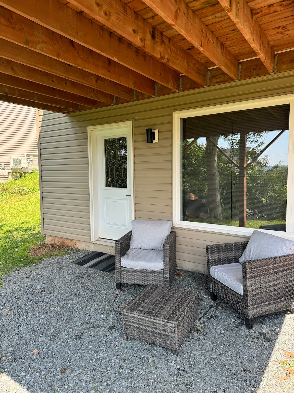 An outdoor seating area is featured with two wicker chairs and a small table, situated under a covered porch. Natural light enters through a large window, revealing a view of the surrounding greenery.