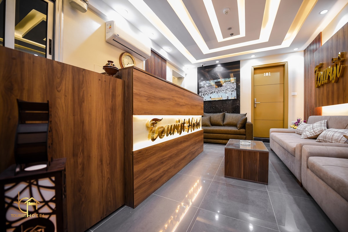 The reception area features a modern wooden desk with illuminated signage. Soft seating is arranged nearby on a glossy tiled floor, complemented by neutral-colored walls and subtle lighting. A contemporary artwork is displayed in the background, enhancing a welcoming environment for guests.