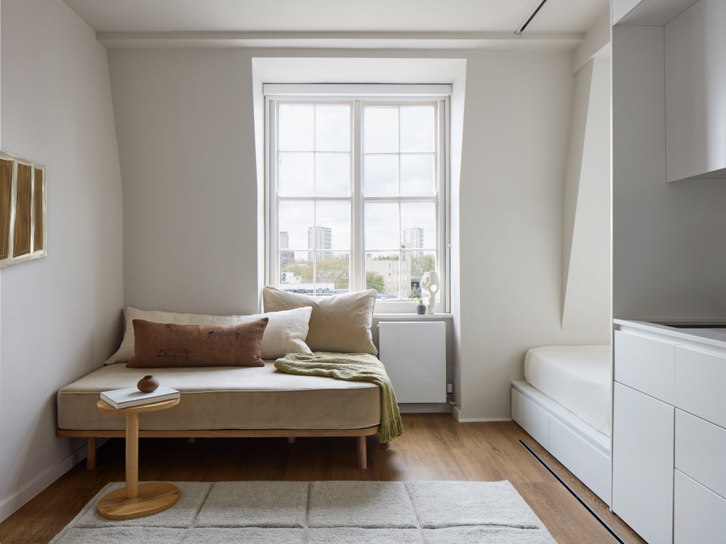 A light-filled space features a cozy seating area with a neutral-colored chaise lounge accompanied by a small wooden side table. A soft throw drapes over the lounge, and large windows allow natural light to illuminate the room, creating a calm atmosphere.