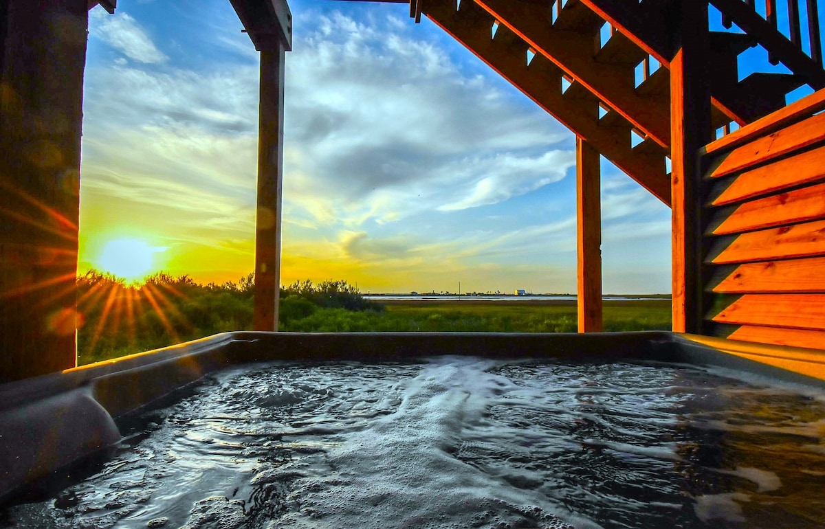 A hot tub is positioned beneath a wooden structure, capturing the serene evening sky and a setting sun. Soft waves ripple across the water's surface, with greenery visible in the foreground and a distant horizon line marking the bay.
