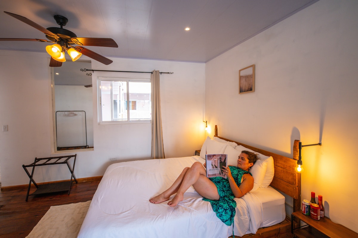 A spacious bedroom is equipped with a queen-sized bed, where a guest is seated comfortably, reading a book. A ceiling fan is visible above, and natural light enters through a window adorned with light curtains. A clothing rack and an accent chair are positioned near the bed.