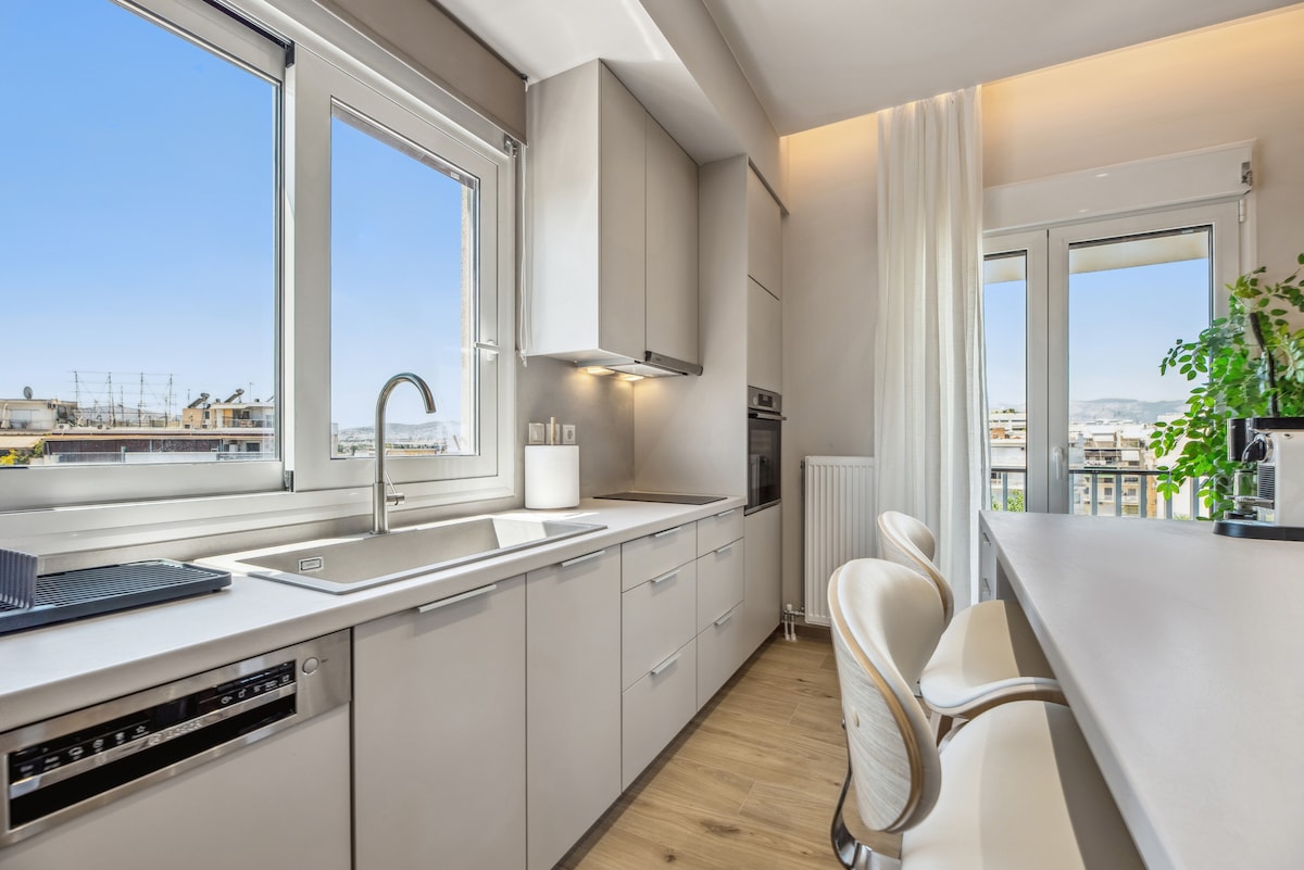 A modern kitchen is presented with sleek cabinetry and a spacious countertop. The area is flooded with natural light through large windows, offering views of the surrounding city. Bar stools are positioned at the counter, providing a casual dining option.