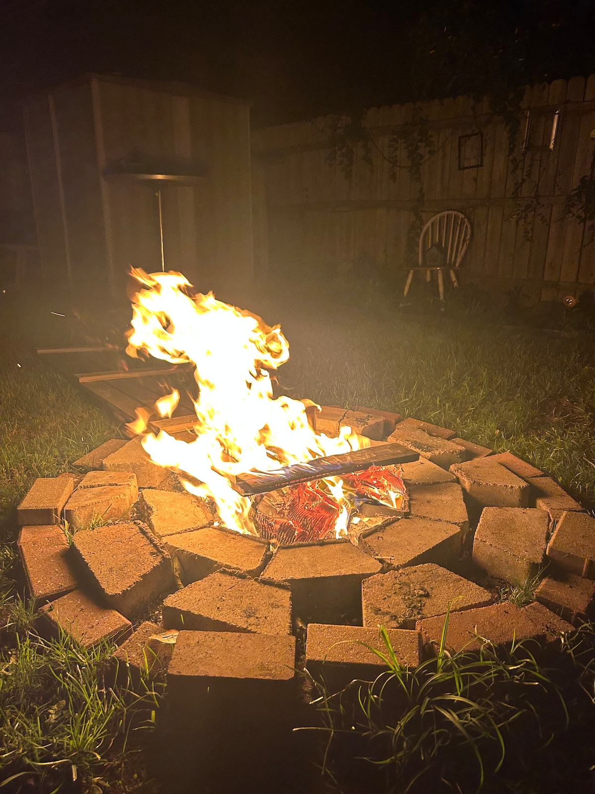 A circular fire pit constructed from bricks is filled with a lively flame, casting a warm glow in the dark outdoor space. The surrounding grass is visible, and a chair is positioned nearby in the background, adding to the inviting atmosphere.