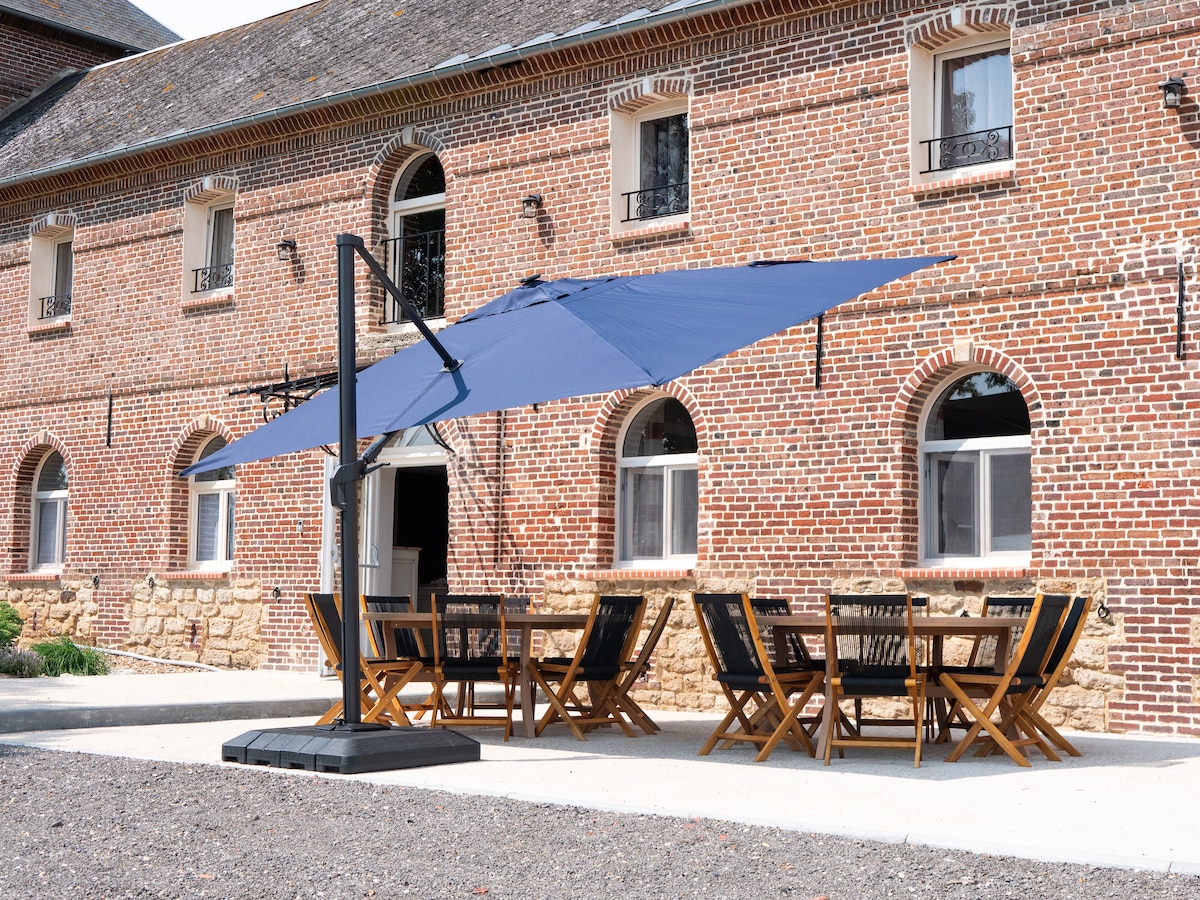 An outdoor dining area is presented, featuring a large table surrounded by multiple wooden chairs. A large navy umbrella provides shade. The rustic brick building serves as a backdrop, showcasing arched windows and a blend of natural stone and brickwork.